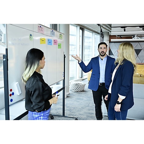 Trois personnes se tiennent devant un tableau blanc. Un homme gesticule pour expliquer. Des post-it sur le tableau.